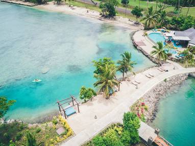 Een helderblauwe zee met palmbonen en een wit strandpad met een ligbed bij Koro Sun Resort in Fiji.