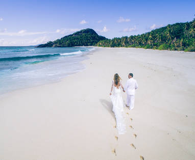 Twee mensen in witte kleding die hand in hand lopen over een wit strand met oceaan en groen bos. 