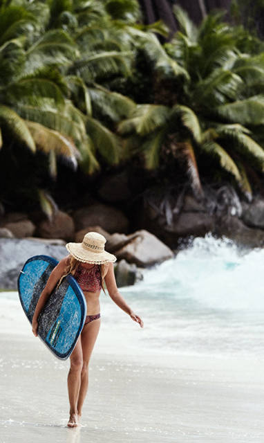 Een zomerse vrouw met een hoedje op en een surfbord met op de achtergrond de oceaan en palmbomen. 