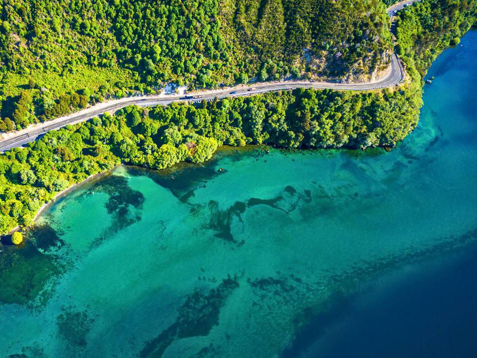 Lake Taupo vanaf boven met de groene heuvels en lichtblauwe oceaan en een autoweg. 