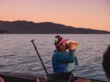 Vrouw die door een schelp blaast vanaf een boot met op de achtergrond de ondergaande zon in het Abel Tasman National Park. 