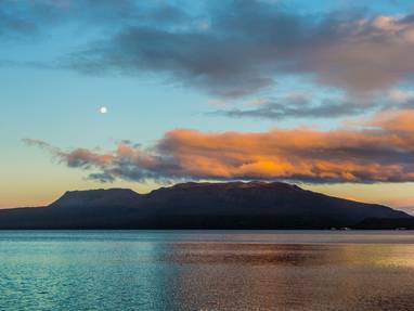 Een meer met donkergekleurde bergen en een geelroze lucht bij Lake Tarawera in Nieuw-Zeeland. 