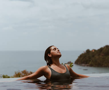 Een dame in een infinity pool met haar ogen dicht en armen over de rand heen met op de achtergrond de oceaan en een baai.