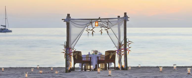 Een tafel op het strand omringd door kaarslicht met als achtergrond een gele lucht en ondergaande zon.