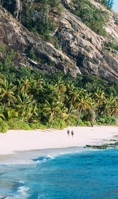Twee mensen die lopen op een wit strand met daarachter palmbomen. 