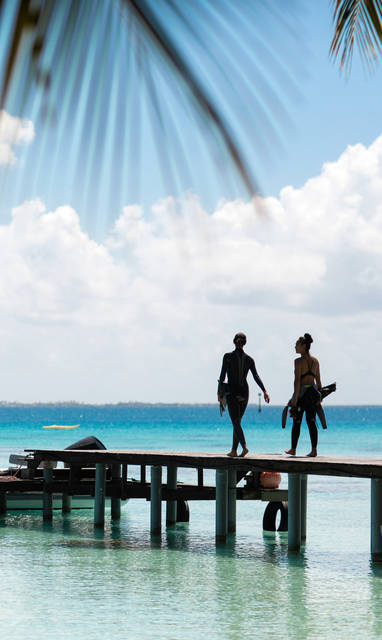 Een boot aangelegd bij een steiger in Frans-Polynesië en twee dames die over de stijger lopen. 