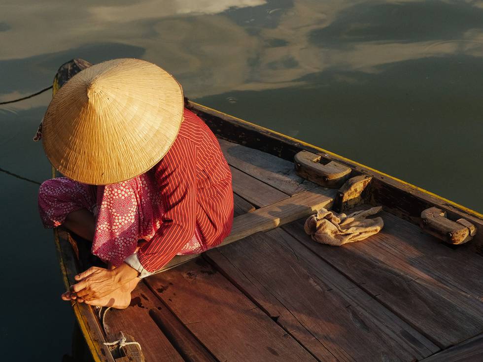 Een persoon met een Aziatische rieten hoed die op een houten boot zit. 